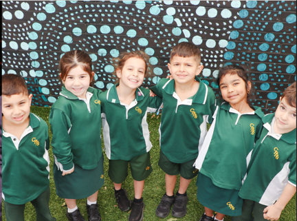 a groups of 6 kindergarten students smiling in front of an Aboriginal mural.
