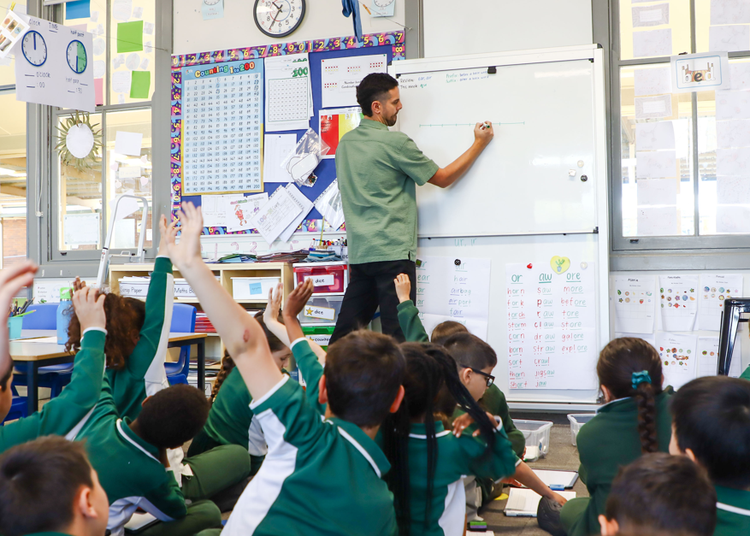 Teacher at the whiteboard in a Stage 1 classroom with students on the floor responding.